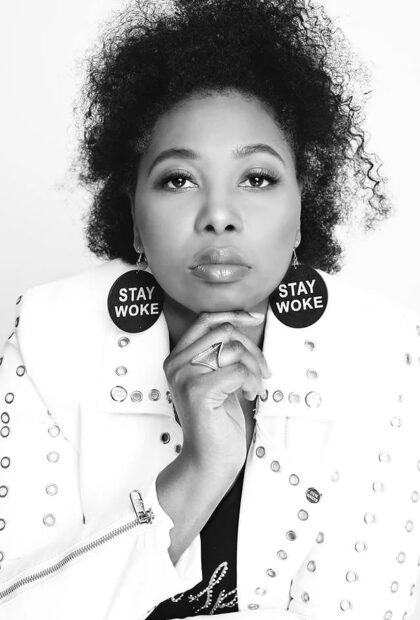 A black and white photograph of Deborah Mouton. She holds her hand under her chin and looks directly at the camera with a serious expression. She wears large circle earrings with text that reads, "Stay Woke."