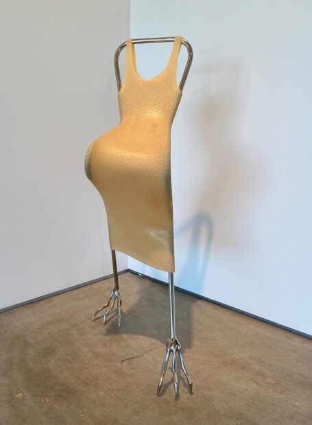 A photograph of a sculpture by Hannah Levy. The sculpture is of a nickel-plated steel form that curves and stands on what looks like bird talons. Stretched over the form is a beige colored silicone object that resembles a sleeveless dress.