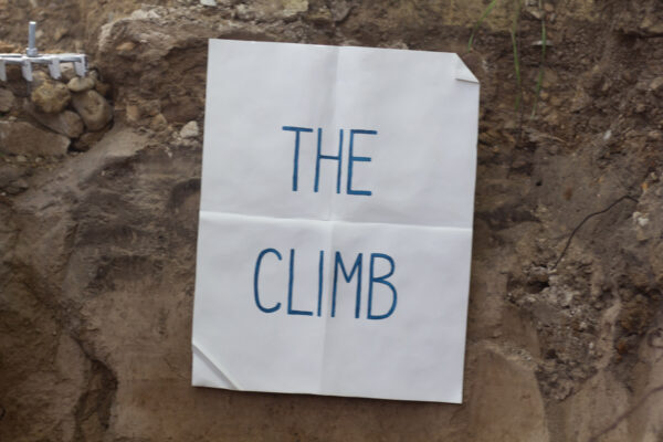 A white piece of paper with perpendicular creases is hung against a wall of raw dirt with the message "The Climb" written on it