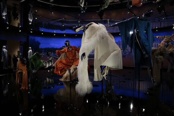 A mannequin wearing a cream-colored dress designed by Hubert de Givenchy, floats in mid-air with its arms dramatically back, as if it had just been hit by another mannequin.