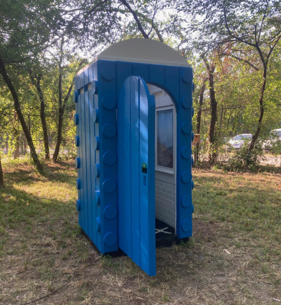A blue custom-built wooden porta potty sits on a grassy field with the door ajar