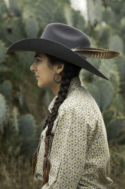 A photograph by Mari Hernandez of Silia Lopez. Lopez is depicted in a profile stance and wears a gray cowboy hat with a feather sticking out. She has long braids and wears a a collared long sleeve shirt with a small pattern.