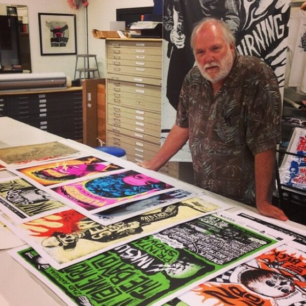 Peter Briggs stands in front of a table with an array of colored prints at Burning Bones Press.