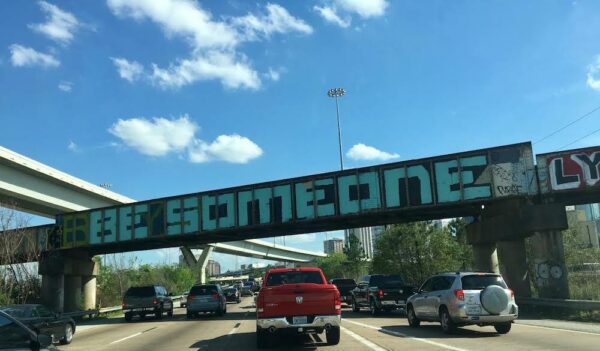 A photo taken from a center lane of I-45 southbound with an overhead train bridge that reads "Be Someone" graffitied in light blue along the side of the bridge.