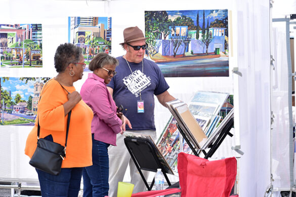 A photograph of a man talking with two women about artistic prints at an open air market.