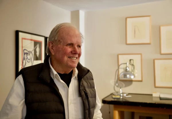 A photograph of Robert Arber in his studio. He wears a black puffy vest over a white button up shirt. There are small framed artworks hanging on the wall behind him.