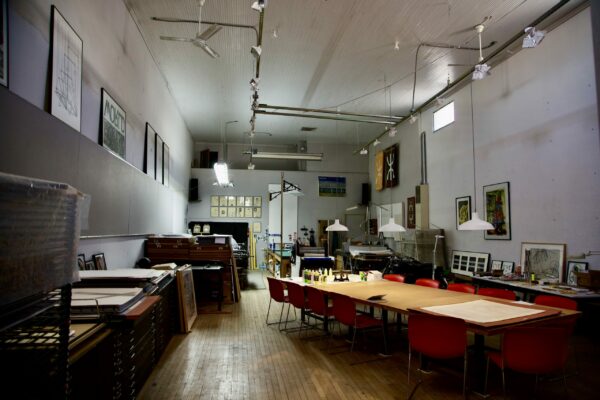 A photograph of Robert Arber's print studio. Flat file storage line the left wall of the studio and there is a long rectangular table with ten red chairs around it. 