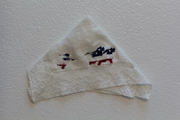 A white laced handkerchief is folded into a triangle and pinned to the wall, with a graphic of a family and small child fleeing on foot is sewn to the handkerchief in a U.S. flag pattern