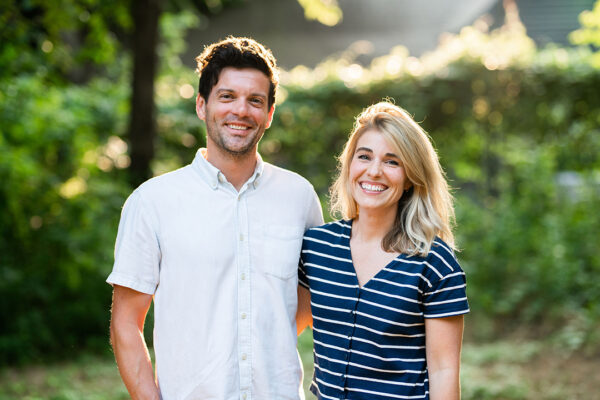 A color photograph of a man and woman standing close together with an arm around each other. They stand in an outdoor setting and both look directly at the camera smiling.