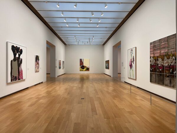 A photograph showing a wood floored, white walled gallery. There are figurative artworks hung along the gallery's walls.