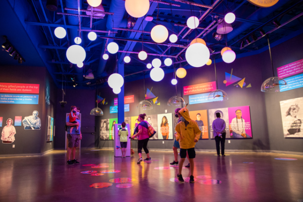 A color photograph of an installation at the DoSeum children's museum. A crowd of people stand under lighted orbs that hang from the ceiling.