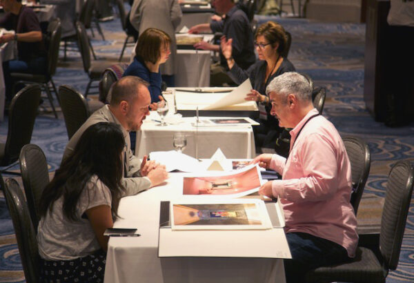 A photograph of small groups of people sitting at tables and reviewing photographic prints.