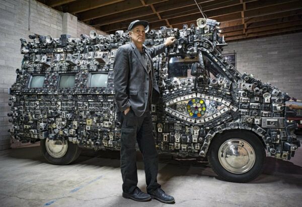 A photograph by Steve Plattner featuring a man standing next to a small van covered in cameras.