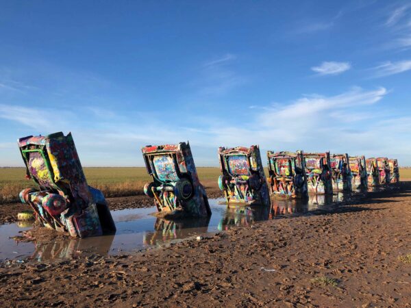 A photograph of the sculpture Cadillac Ranch, which is ten cadillac cars buried, hood first, in the ground. The cars are covered in graffiti.