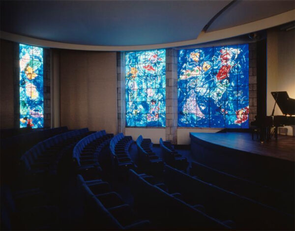 A photograph of the inside of a darkened auditorium. Three large abstract stained-glass panels let in a flood of blue light.