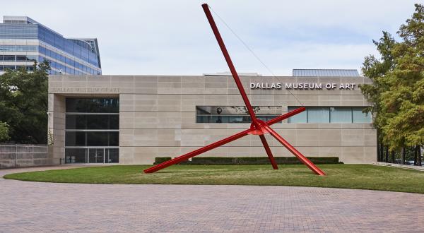 A color photograph of the Ross Street entrance of the Dallas Museum of Art. A large abstract red sculpture stands in a grassy area to the right of the museum entrance.