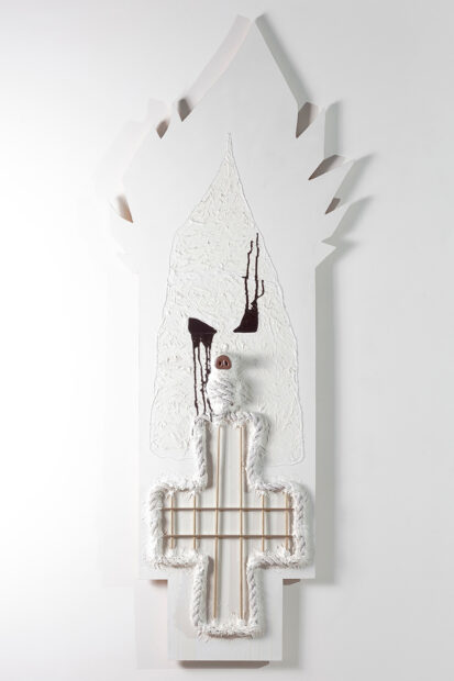 A vertical mixed-media work on a wooden panel by David-Jeremiah. The artwork uses rope and plastic to create shapes and patterns and is mostly covered in white enamel paint. Though simplified, the design seems to reference a KKK hooded figure with a cross.