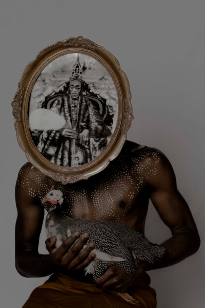 A photograph by Hakeem Adewumi of a seated and shirtless Black man holding a chicken. The man's chest and shoulders are filled with dots of white paint and a framed artwork covers his face. 