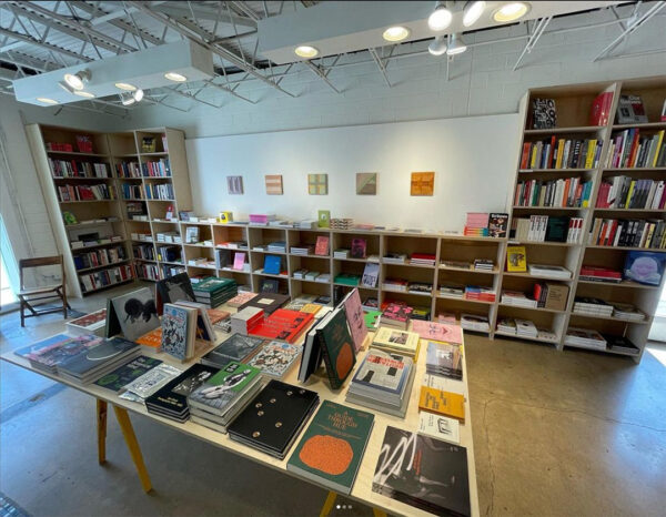 A photograph of the interior of Basket Books & Art. The photograph shows a tabletop covered with books in the middle of the room and a mix of tall and short bookshelves lining the walls.