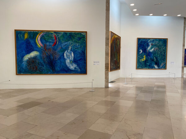 An installation view of the main gallery at the Marc Chagall National Museum. The image shows three works hung on angled walls. The works use mostly shades of blue and green and feature floating figures.