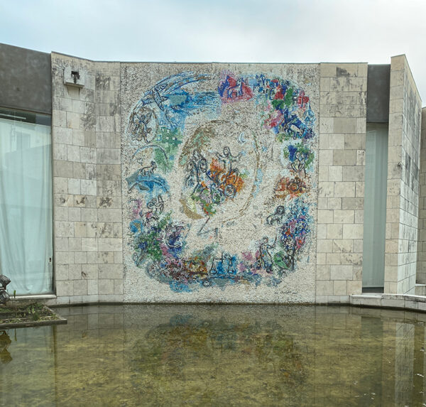 A photograph of a large-scale mosaic featuring the prophet Elijah ascending to the heavens in a chariot of fire. There is a large reflecting pond below the mosaic.