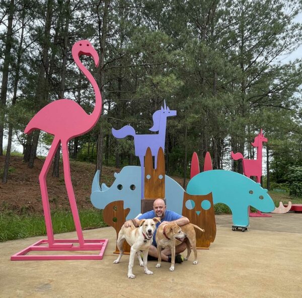 Artist Jeffie Brewer with his two dogs in front of his colorful animal shaped sculptures