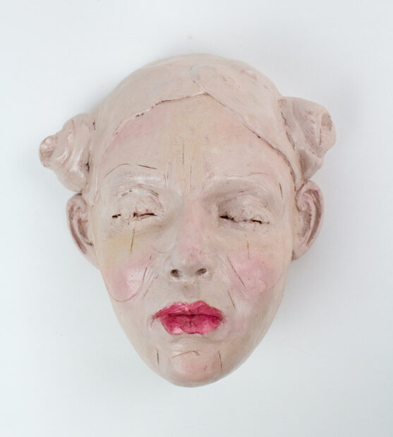 A photograph of a sculpture by Payton Koranek. The sculpture is of a female face with closed eyes and red lipstick. The face and hair, which is arranged in two buns atop the head, are painted light pink. 