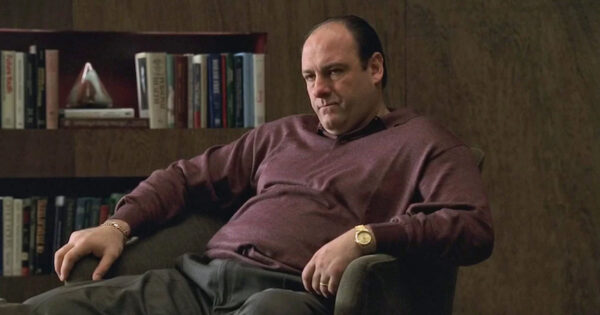 A photograph of Tony Soprano, from 'The Sopranos,' sitting in a chair. He is balding, wearing a sweater, pants, and a wristwatch.