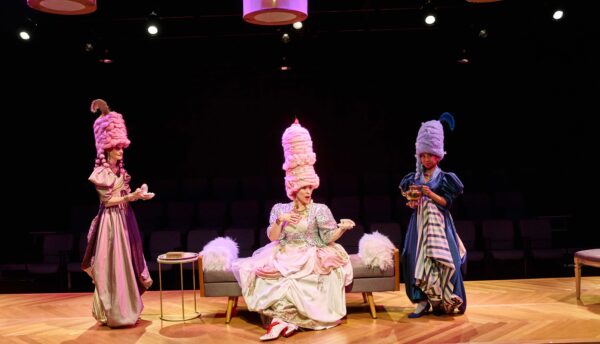 Photo of three women acting on a stage wearing elaborate costumes with tall headpieces