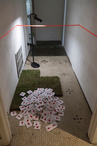 An installation view of the interior hallway of Beefhaus in Dallas. Squares of sod are placed on the ground, and sticky note pads with the Google Maps pin printed on them are shuffled into a pile on top of the sod.