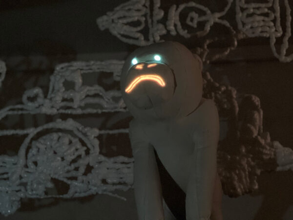  A dark photograph of a fabric sculpture of a monkey with lights that illuminate its eyes, nose, and mouth.