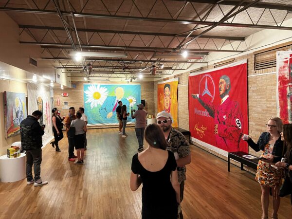 Photo of an exhibition opening with large scale paintings hanging in front of a brick wall