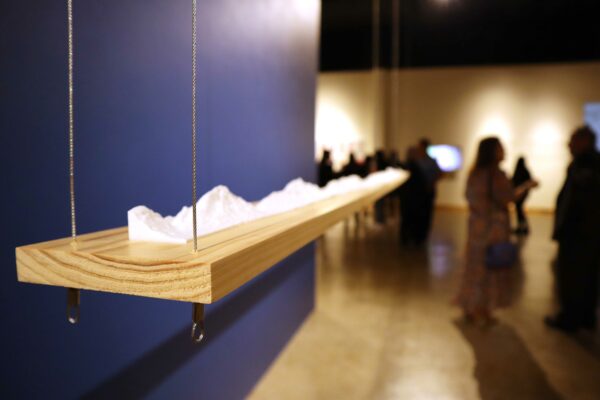 Image of a detail of a three dimensional work suspended agains a blue wall during the opening of an exhibition
