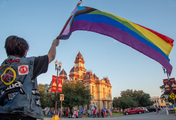 A photograph by Ursula Rogers of a person waving a large rainbow flag. The person is in the foreground of the photograph and has short hair and wears a jean vest with patches on the back. In the distance, across the street and near a courthouse is a crowd of people waving U.S. and Texas flags.