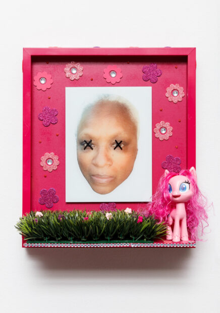 A mixed-media work by Vicki Meek. The artwork features a photograph of the artist's face with two black Xs over her eyes. The photograph is presented on a pink frame with pink flower stickers. A small shelf extends from the base of the work and holds a small patch of fake grass and a pink My Little Pony toy.