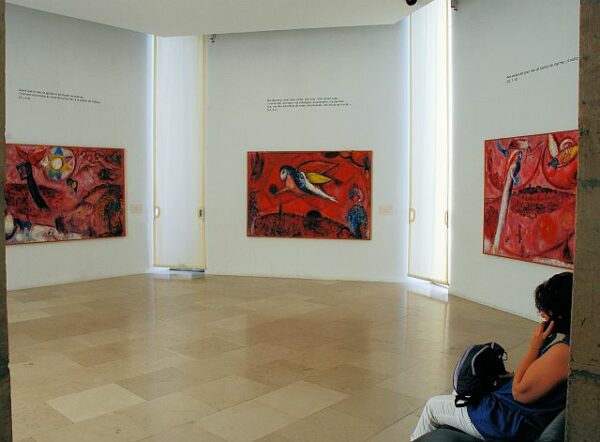 An photograph showing an installation of three paintings by Marc Chagall. All three images use shades of red and are part of the artist's "Song of Songs" series.