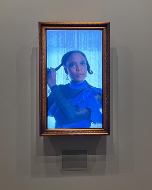 A still image of a video installation by Adam Delphine Fawundu. The video shows a Black woman dressed in blue with her hands positioned to undo her braids. 