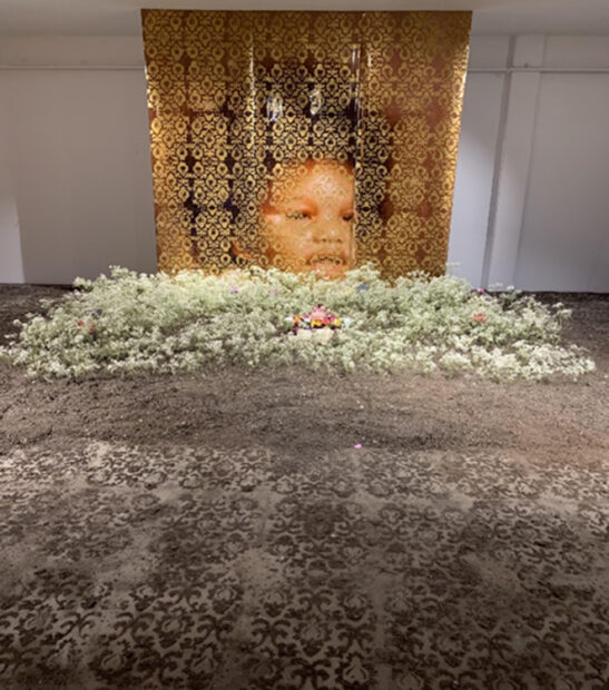 Photograph of an art installation featuring a large portrait with gold patterning overlaid on top of it. The portrait hangs from the ceiling in the background. In the foreground of the scene, it appears dirt is laid out in an ornate pattern. A larger mound of dirt appears in the middle ground of the scene and seems to have short foliage sticking out of it.