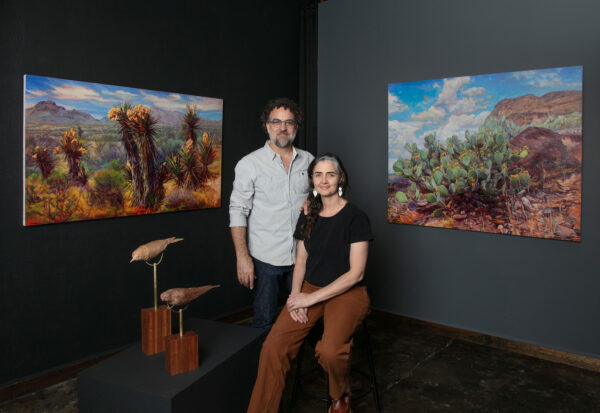 A photographic portrait of Dana Younger and Felice House. Work by both artists flank the couple: desert landscape paintings, and sculptures of bird-looking creatures.