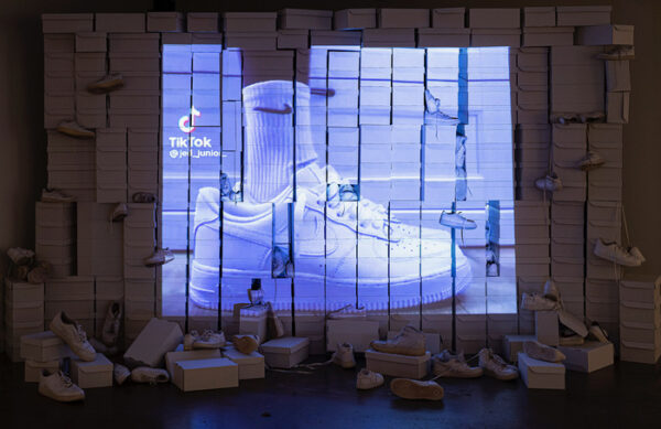 An installation photograph of a work by Ciara Elle Bryant. A video of a person wearing a pair of white Nike Air Force 1 shoes. The video is projected onto a makeshift wall of white shoe boxes with Nike Air Force 1 shoes spread out on the floor and hanging on some of the boxes.