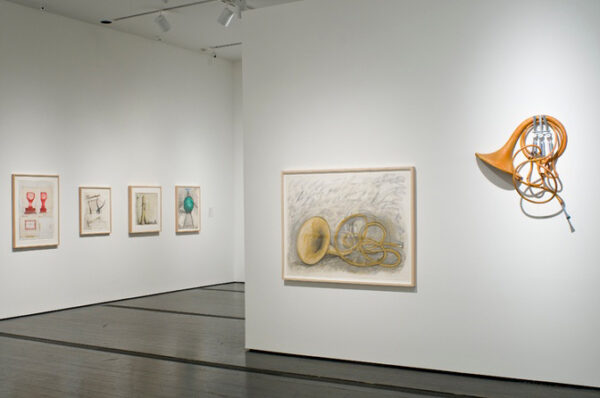 A photograph of a white gallery space with many framed drawings. In the foreground is a drawing of a french horn, hanging on a wall next to a sculpture of a french horn.