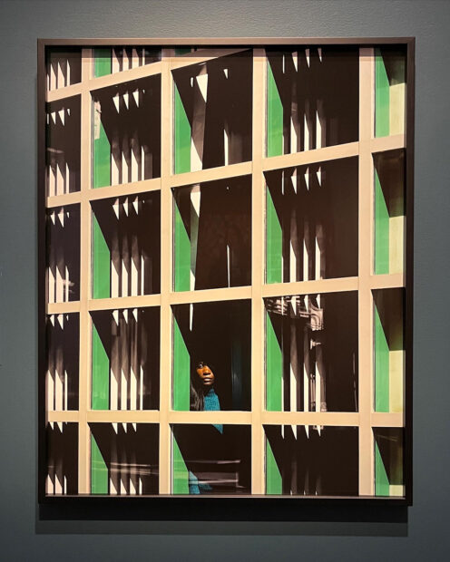 A photograph of a woman peering out of a window. The image is filled with a grid of windows and the shadows create the appearance of green and black right triangles.