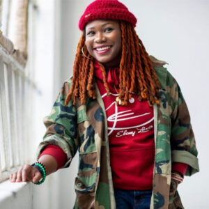 A headshot of Ebony Lewis, she stands next to a window and wears a red hat and a red long sleeve shirt with a camouflage shirt layered on top. 