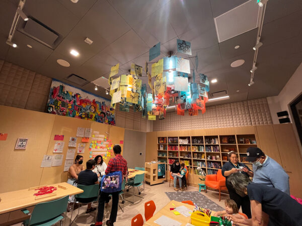 A photograph of visitors engaging with various components of the multipurpose educational space within the Molina Family Latino Gallery. The space has table top activities, a response wall, and a library.