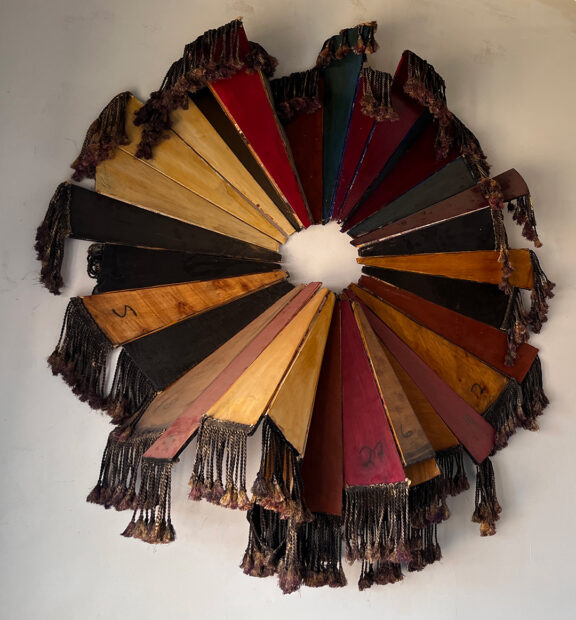 A photograph of a sculptural work by Emily Sloan. The mixed media object hangs on a gallery wall and is made from wood triangular pieces that come together to make a wheel. At the end of each triangle are tassels. 
