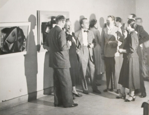 A black and white photograph from 1950 of a crowd of people in a gallery with works by Paul Fontaine hanging on the wall. 