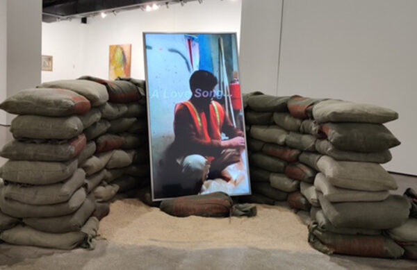 An installation by Goran Maric featuring a video work displayed on a vertically positioned screen which is surrounded by military grade sand bags.