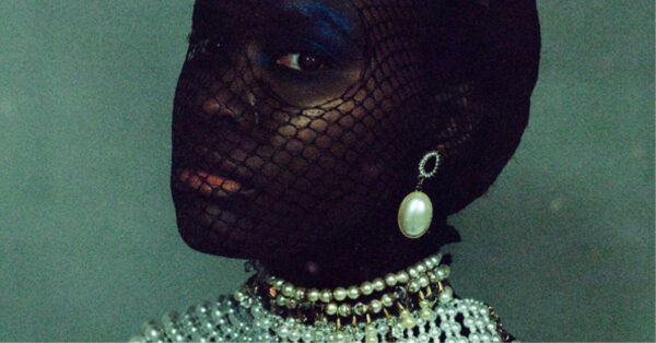A detail of a photograph by Grace Nicole. The image depicts a black woman with a fishnet stocking over her face and large holes cut out around her eyes so that they are unobstructed. She wears a large pearl earring and an elaborate pearl necklace.
