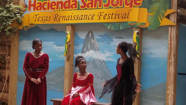 A photograph of a performance at the Texas Renaissance Festival of three Black women dressed in Renaissance era clothing while talking on stage.