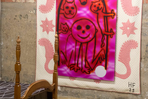 An artwork consisting of a a white blanket embroidered with pink paisley details frames a magenta painting on photosensitive paper with a human figure kneeling beneath and arc of cartoonishly rendered cats. The painting is hung on a rough concrete wall beside a child's bed.